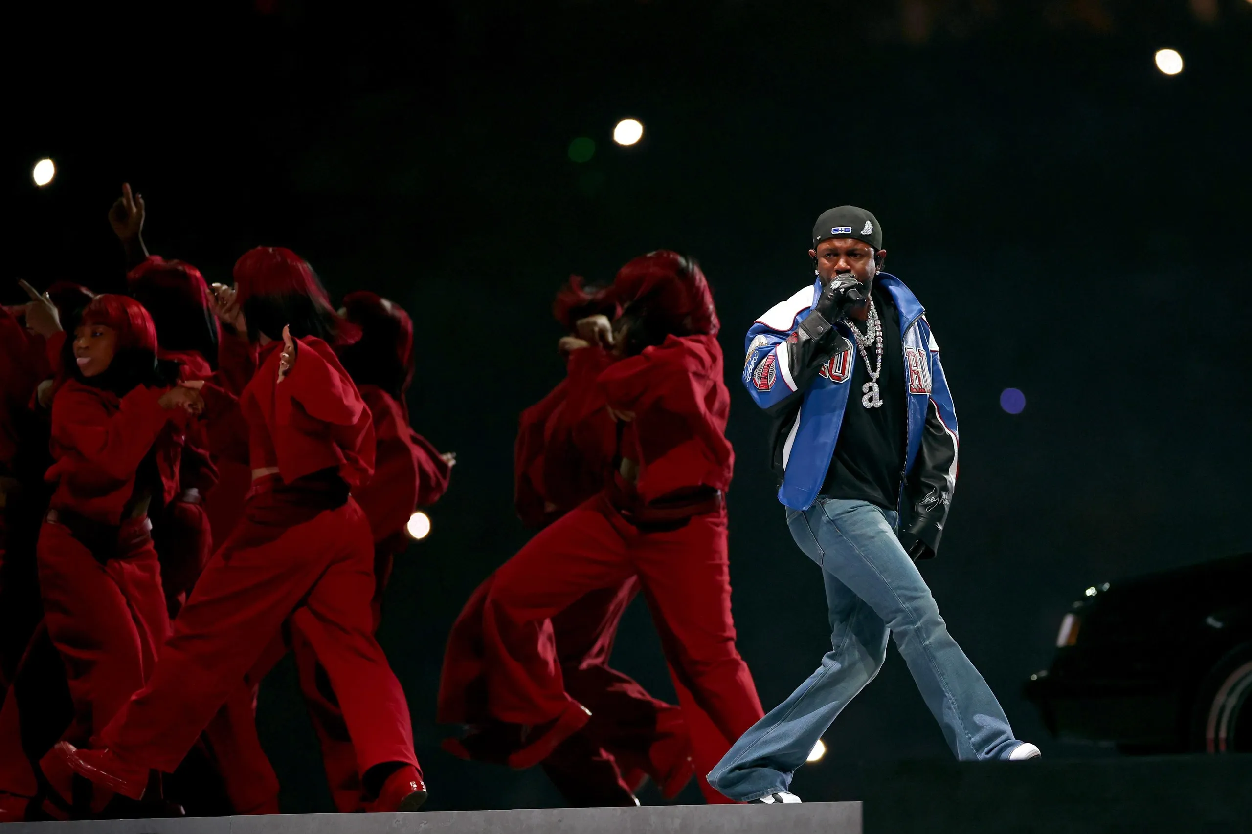 A rapper striding across a stage in jeans and baseball jacket with backing dancers in red behind him