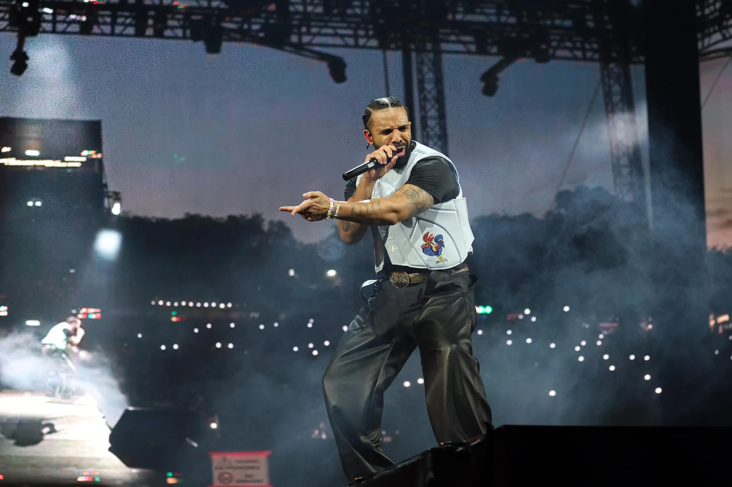 A bearded rapper singing on stage with black T-shirt and white vest and black trousers 
