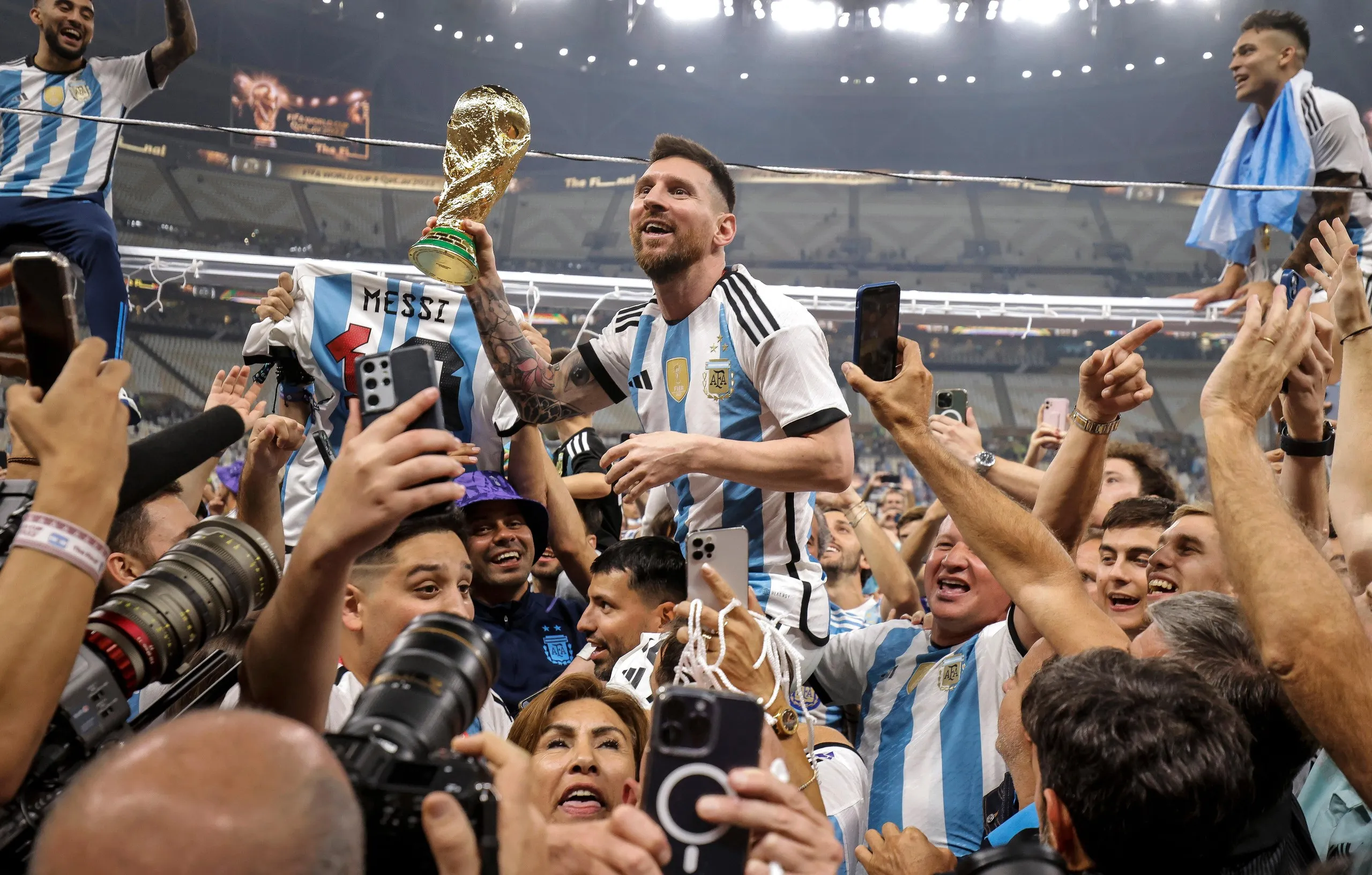 Messi is held aloft by teammates with the World Cup trophy in his hand as he is surrounded by photographers